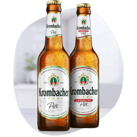 Zwei Krombacher Pils-Flaschen, eine mit alkoholhaltigem und eine mit alkoholfreiem Bier, stehend vor unscharfem Hintergrund.