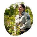 Forstmitarbeiterin in beiger Uniformjacke mit 'Forstverwaltung' Abzeichen sitzt im Wald zwischen grünen Pflanzen