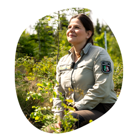 Forstmitarbeiterin in beiger Uniformjacke mit 'Forstverwaltung' Abzeichen sitzt im Wald zwischen grünen Pflanzen
