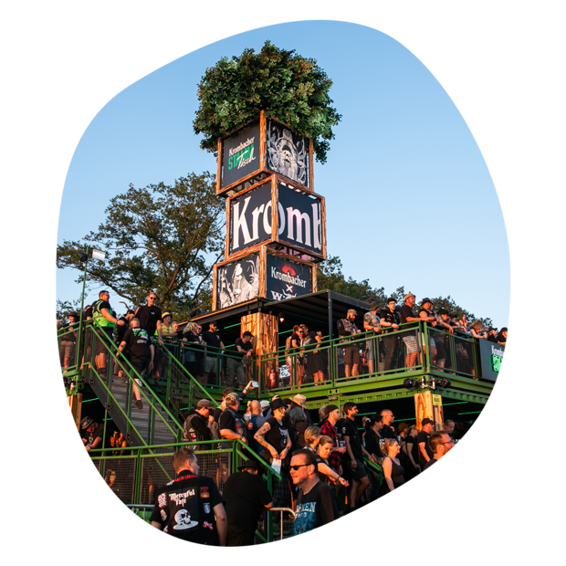 Menschen auf einer mehrstöckigen Festivalbühne mit Krombacher-Werbung und Baum-Dekoration im Hintergrund
