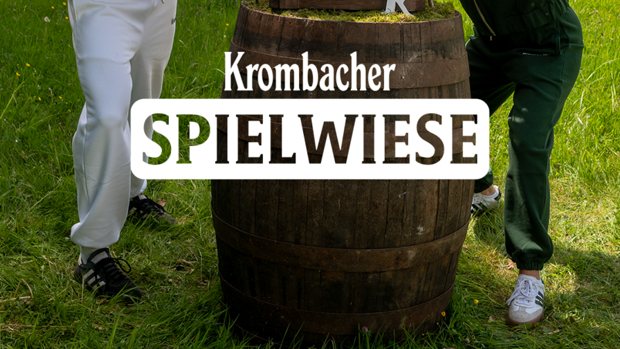 Drei Personen stehen draussen auf einer Wiese um ein Holzfass mit Krombacher-Logo und der Aufschrift „Krombacher Spielwiese“. Zwei von ihnen, in weissem und grünem Trainingsanzug, beugen sich streitlustig gegeneinander, während die Person in goldener Jacke dazwischengeht und überrascht schaut.