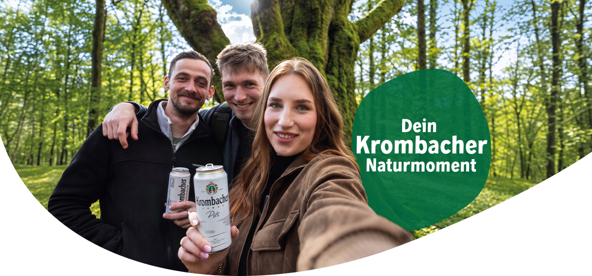 Drei junge Erwachsene machen ein Selfie in einem sonnigen Wald vor einem grossen, moosbewachsenen Baum; zwei halten Getränkedosen mit Krombacher-Logo in der Hand. Rechts im Bild ein gruener Kreis mit dem Text „Dein Krombacher Naturmoment“.