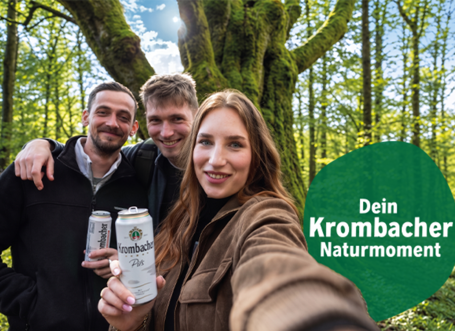 Drei junge Erwachsene machen ein Selfie in einem sonnigen Wald vor einem grossen, moosbewachsenen Baum; zwei halten Getränkedosen mit Krombacher-Logo in der Hand. Rechts im Bild ein gruener Kreis mit dem Text „Dein Krombacher Naturmoment“.