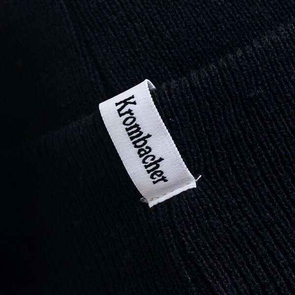 Schwarze Beanie mit weißem Etikett und schwarzem Schriftzug 'Krombacher'.