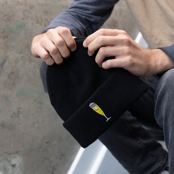 Person hält schwarze Beanie mit gesticktem Bierglas-Logo in der Hand.