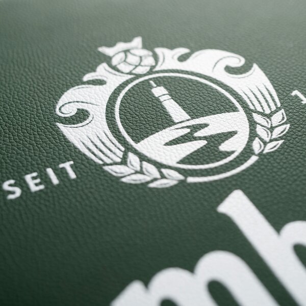 Weißes Krombacher-Logo mit Turm, Hopfen und Gerstenähren auf grünem, strukturiertem Hintergrund, teilweise sichtbarer Schriftzug 'SEIT'.