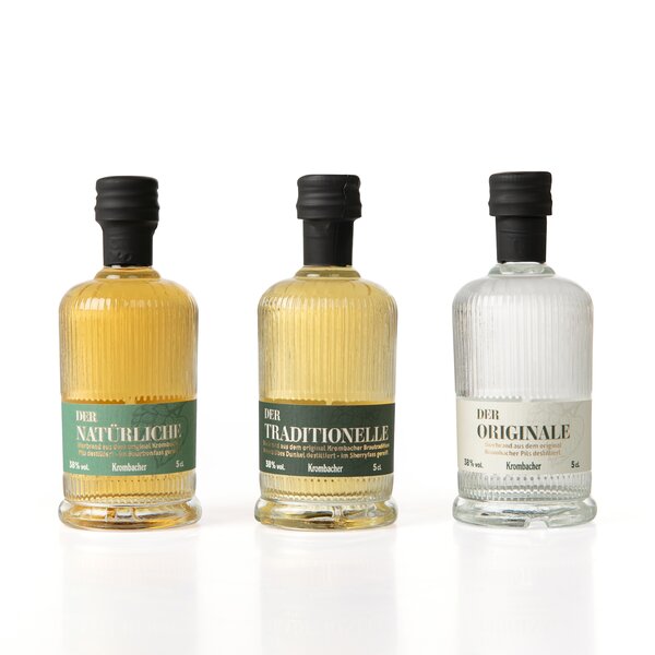 Drei kleine Krombacher Probierbrand-Flaschen mit schwarzen Verschlüssen, beschriftet als 'Der Natürliche', 'Der Traditionelle' und 'Der Originale'.