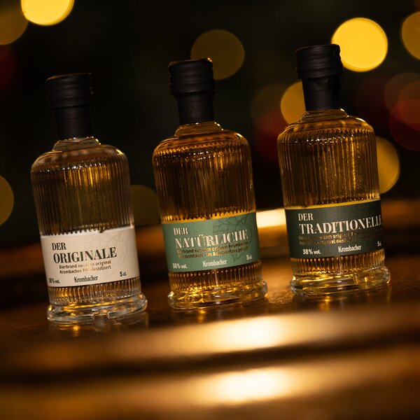 Drei kleine Krombacher Spirituosenflaschen mit schwarzen Verschlüssen, beschriftet mit 'Der Originale', 'Der Natürliche' und 'Der Traditionelle', vor unscharfem Lichthintergrund.