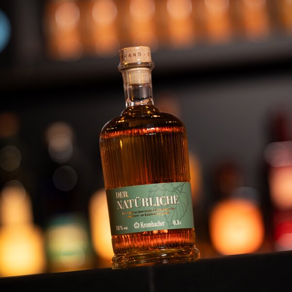 Flasche Krombacher Bierbrand 'Der Natürliche' mit grünem Etikett, 0,5 Liter, 38 % Vol., bernsteinfarbener Inhalt, auf dunklem Untergrund.