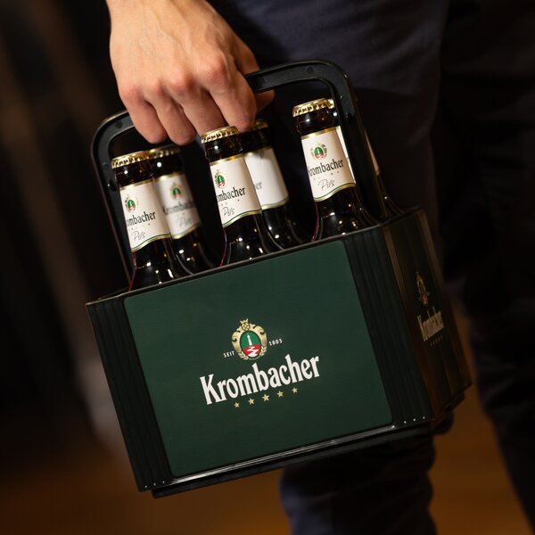 Hand hält einen dunkel grünen Krombacher-Sechserträger mit grüner Front und sechs Flaschen Krombacher Pils.