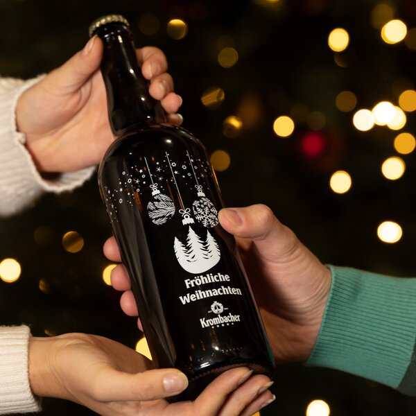 Zwei Hände halten eine braune Krombacher-Flasche mit weißen Weihnachtsmotiven und dem Text 'Fröhliche Weihnachten'.