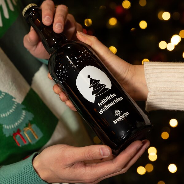 Zwei Hände halten eine braune Krombacher-Flasche mit weißem Druck, darauf ein stilisierter Weihnachtsbaum und der Text 'Fröhliche Weihnachten'.