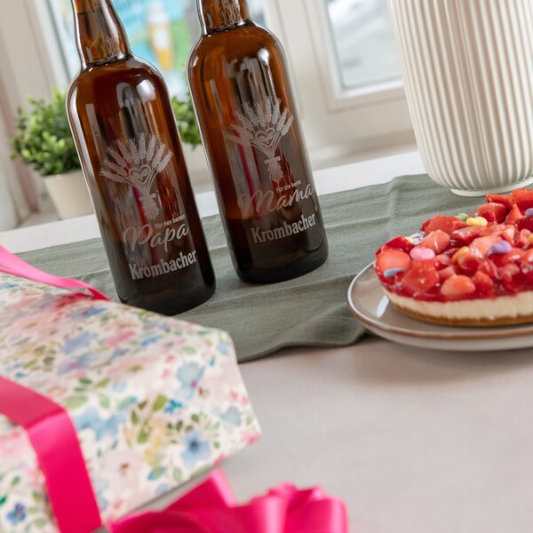 Zwei braune Krombacher-Flaschen mit Gravur "Für den besten Papa" und "Für die beste Mama" auf einem Tischläufer neben einem Erdbeerkuchen und einem Geschenk mit pinkem Band.