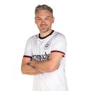 Mann mit verschränkten Armen trägt weißes Trikot mit schwarzem, roten und goldenen Streifen an den Ärmeln und aufgedrucktem Krombacher Logo.