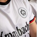 Ausschnitt eines weißen Trikots mit schwarzem Kragen, einem Wappen auf der Brust und dem Schriftzug 'Krombacher' sowie einem Ärmel mit schwarz-rot-goldenem Rand